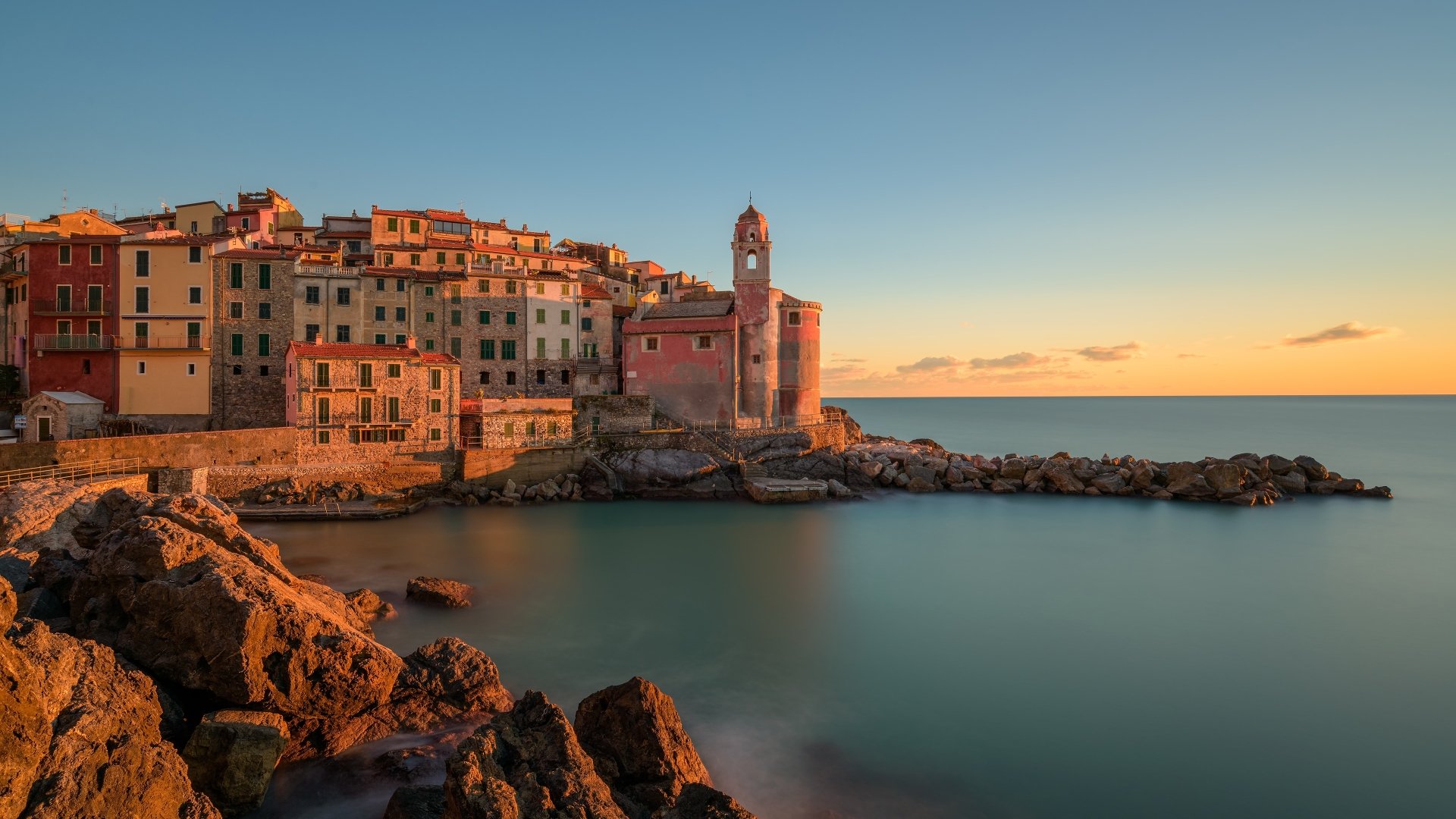 4K Ultra HD PC desktop wallpaper: Tellaro village on a rocky coast, man-made pastel buildings and church tower bathed in sunset light above the calm sea.