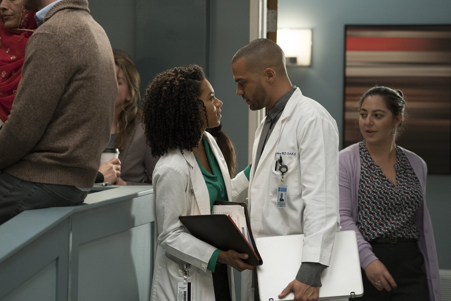 A scene from Grey's Anatomy featuring two doctors in a moment of connection, with other characters in the background, set against a modern hospital interior. HD desktop wallpaper.