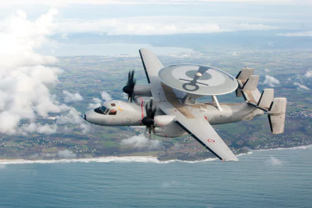 HD desktop wallpaper featuring a military Northrop Grumman E-2 Hawkeye warplane flying over coastline with ocean and distant land under a partly cloudy sky.