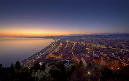 A stunning view of Nice, France at twilight, showcasing the vibrant city lights along the coast as the sun sets over the calm Mediterranean Sea.