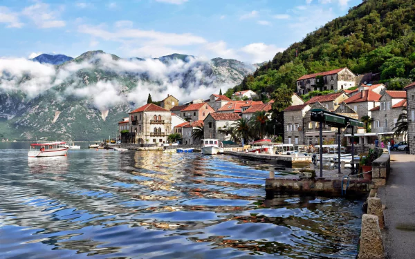 HD desktop wallpaper of the man-made village of Perast in Montenegro, featuring waterfront houses, boats, and lush green hills under a partly cloudy sky.