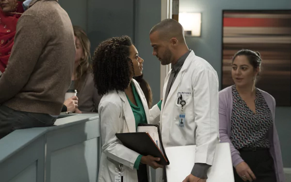 A scene from Grey's Anatomy featuring two doctors in a moment of connection, with other characters in the background, set against a modern hospital interior. HD desktop wallpaper.