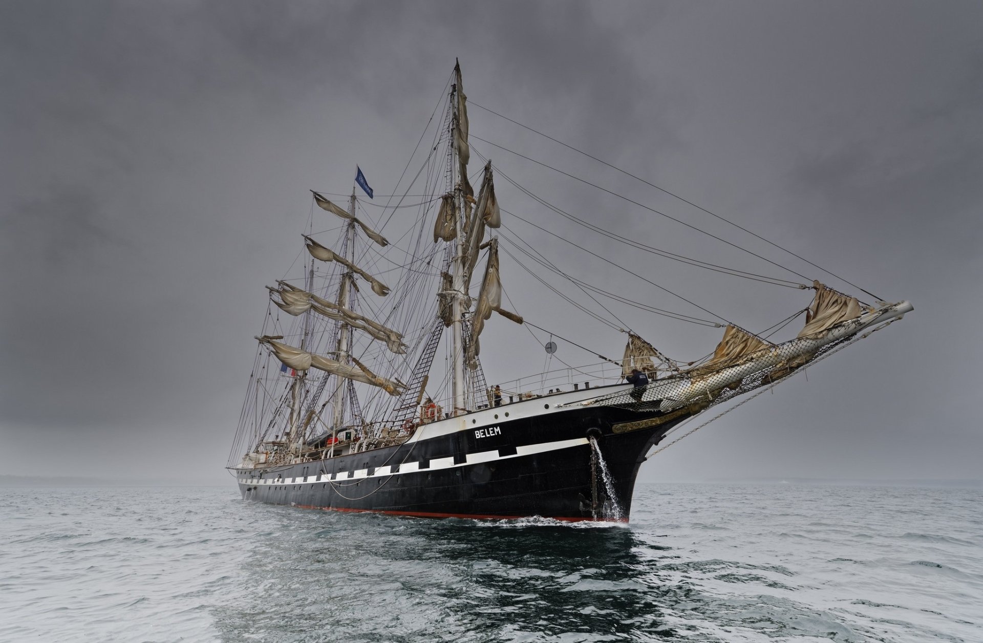 HD PC desktop wallpaper of a majestic barque sailboat vehicle with full masts gliding on calm gray seas beneath an overcast sky.