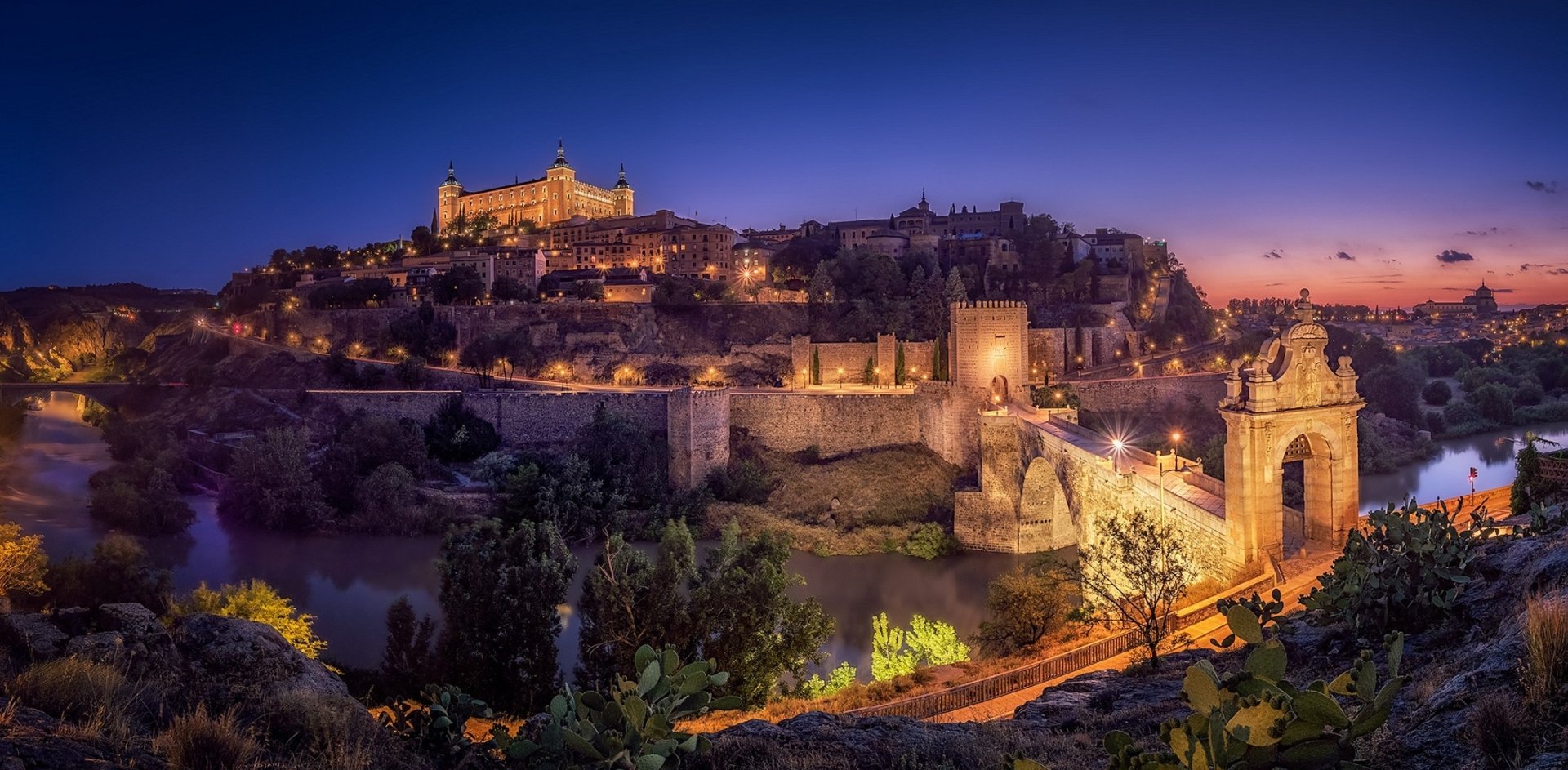 Night view of the historic man-made bridge and buildings in Toledo, Spain, illuminated against a deep blue sky in this HD desktop wallpaper.