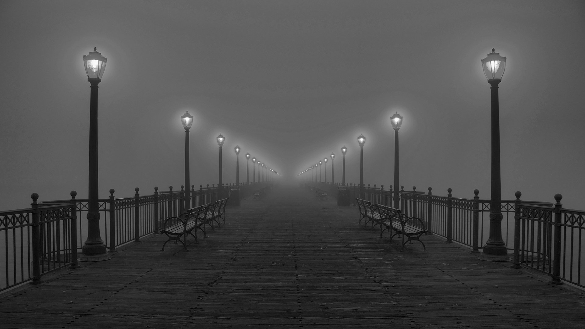 A misty pier illuminated by evenly spaced lamps with benches along the sides, captured in a high-definition black-and-white desktop wallpaper.