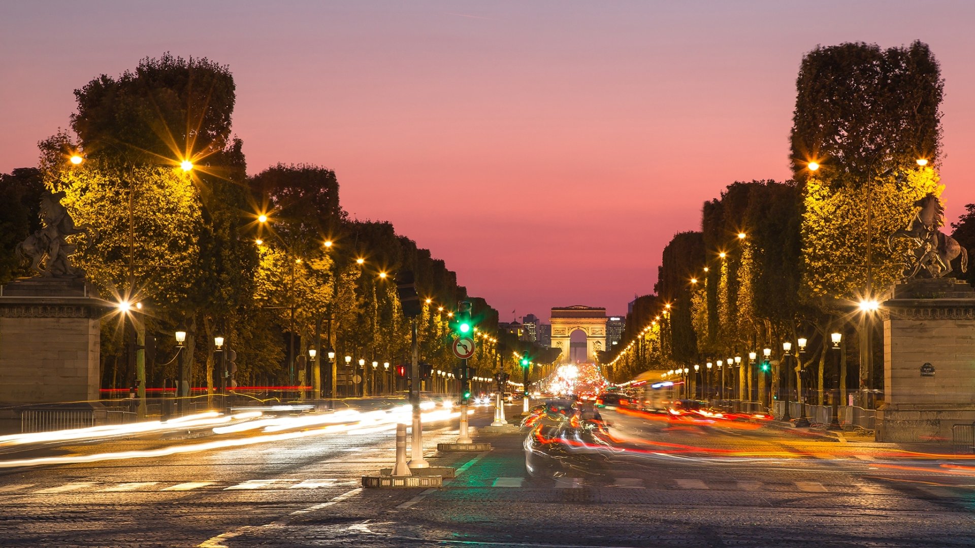 Time-lapse night view of the Champs-Élysées in Paris with illuminated trees and the Arc de Triomphe in the background as a vibrant HD desktop wallpaper.