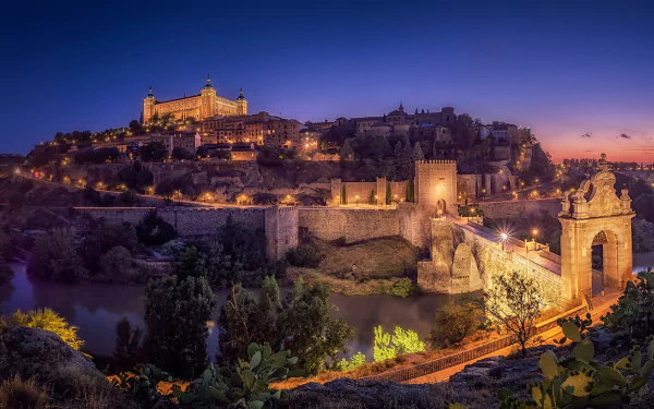 Night view of the historic man-made bridge and buildings in Toledo, Spain, illuminated against a deep blue sky in this HD desktop wallpaper.