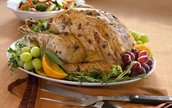 HD desktop wallpaper featuring a roasted chicken garnished with fresh grapes, sliced oranges, and herbs, accompanied by a carving knife and fork on a table setting.