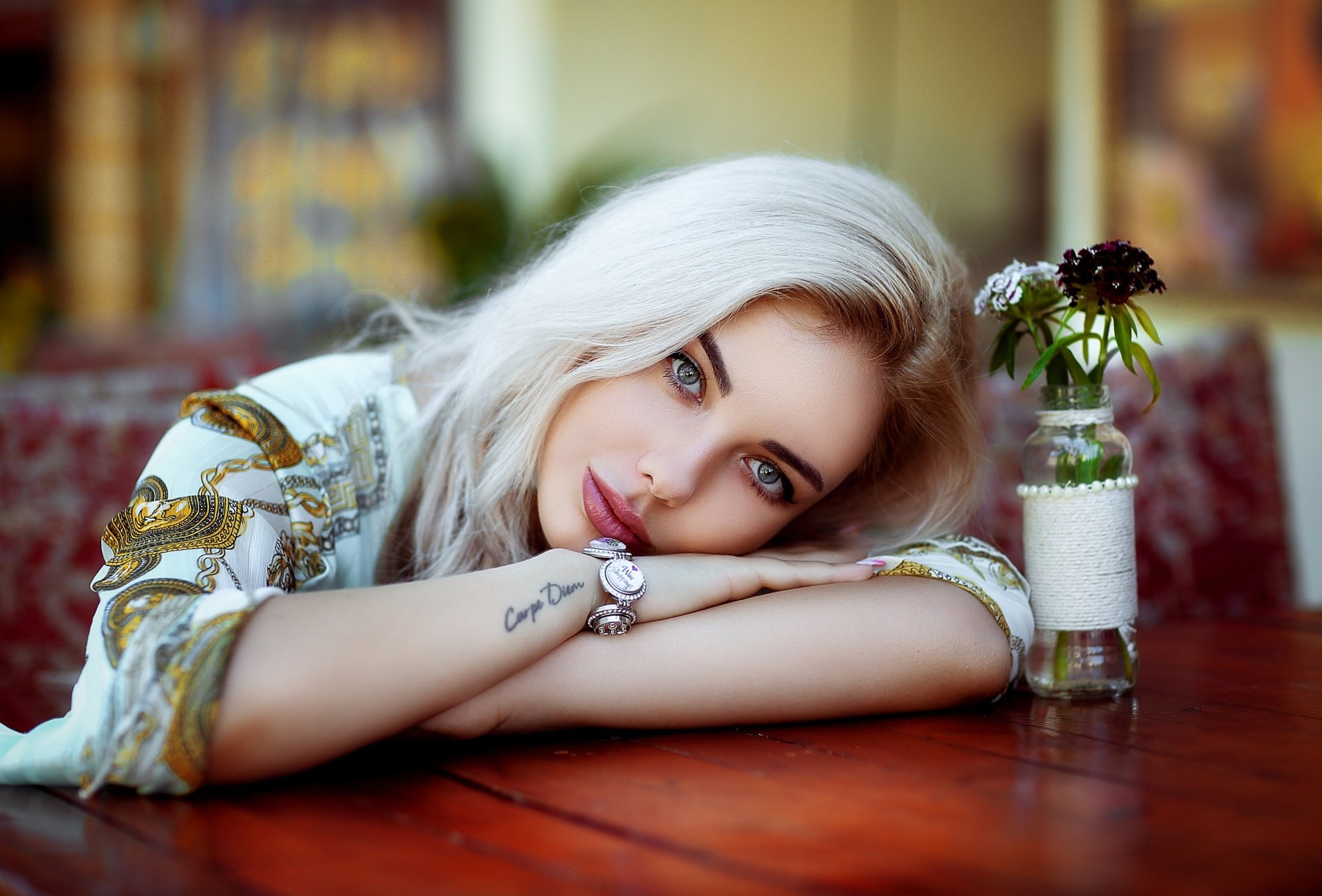 HD desktop wallpaper featuring a blonde woman model resting on a wooden table with a small flower vase, captured by Hakan Erenler.