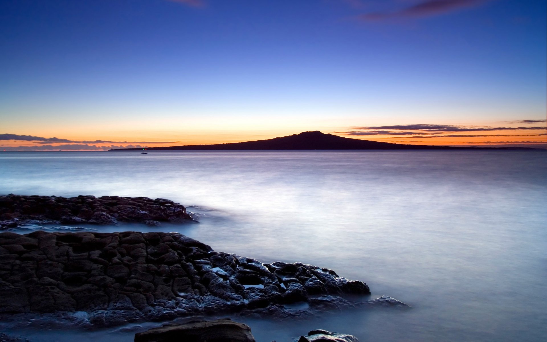 A serene sunrise over a tranquil ocean, with soft clouds framing a distant mountain, rocky coastlines, and a calm beach, creating a stunning HD nature wallpaper.