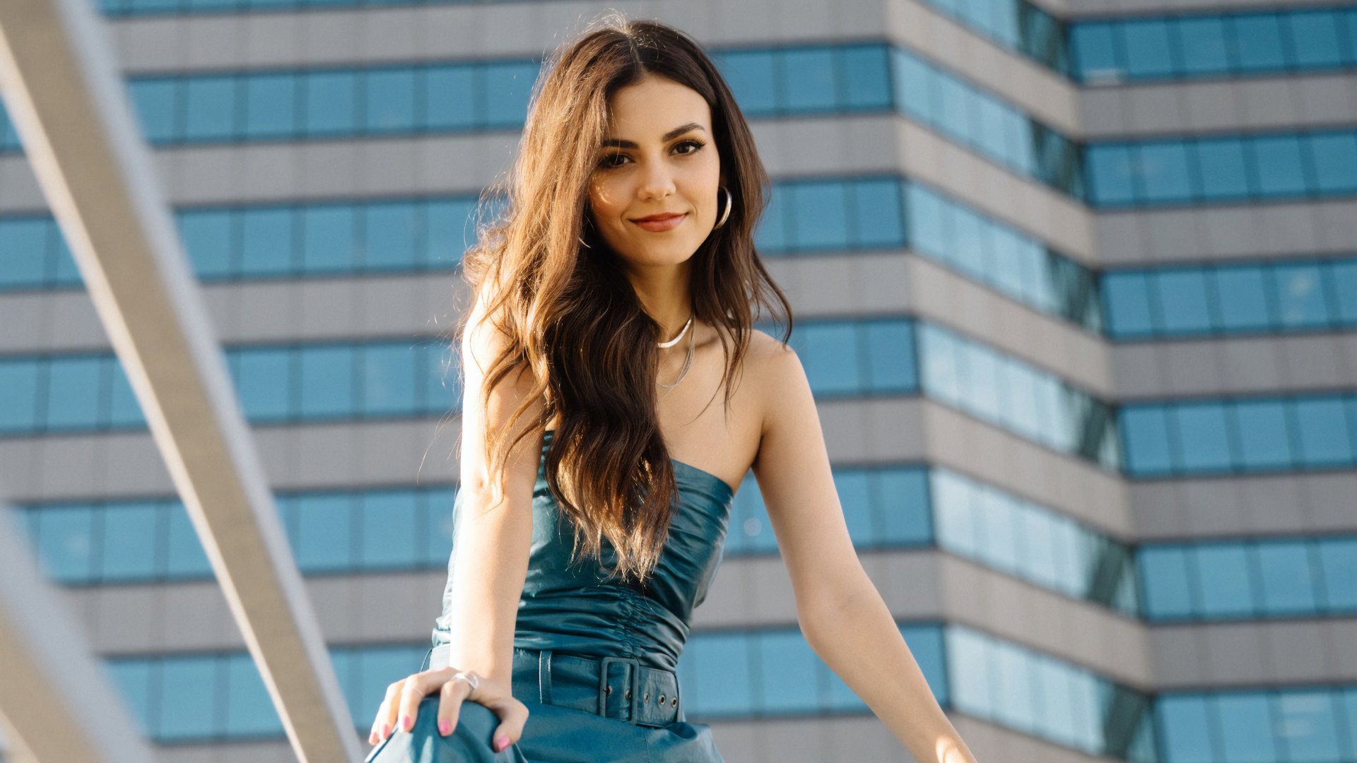 Long-haired brunette woman in a teal dress posing in front of a modern glass high-rise — 4K Ultra HD desktop wallpaper.