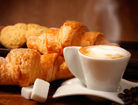 A steaming cup of coffee with frothy milk, sugar cubes, and golden croissants on a warm-toned background, captured in 4K Ultra HD for a PC desktop wallpaper.