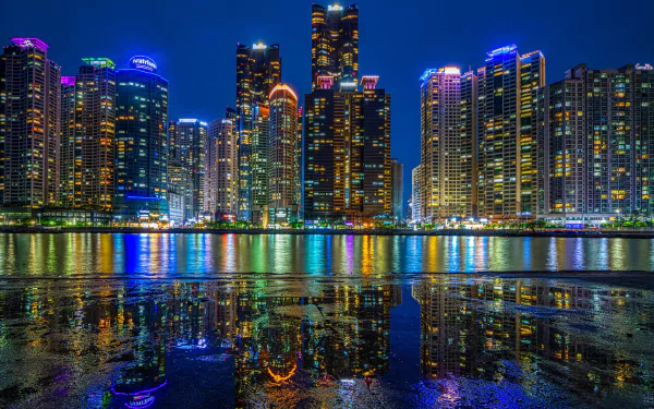 Night view of Busan’s illuminated skyscrapers reflecting on water, showcasing South Korea’s vibrant cityscape with colorful lights and towering buildings.