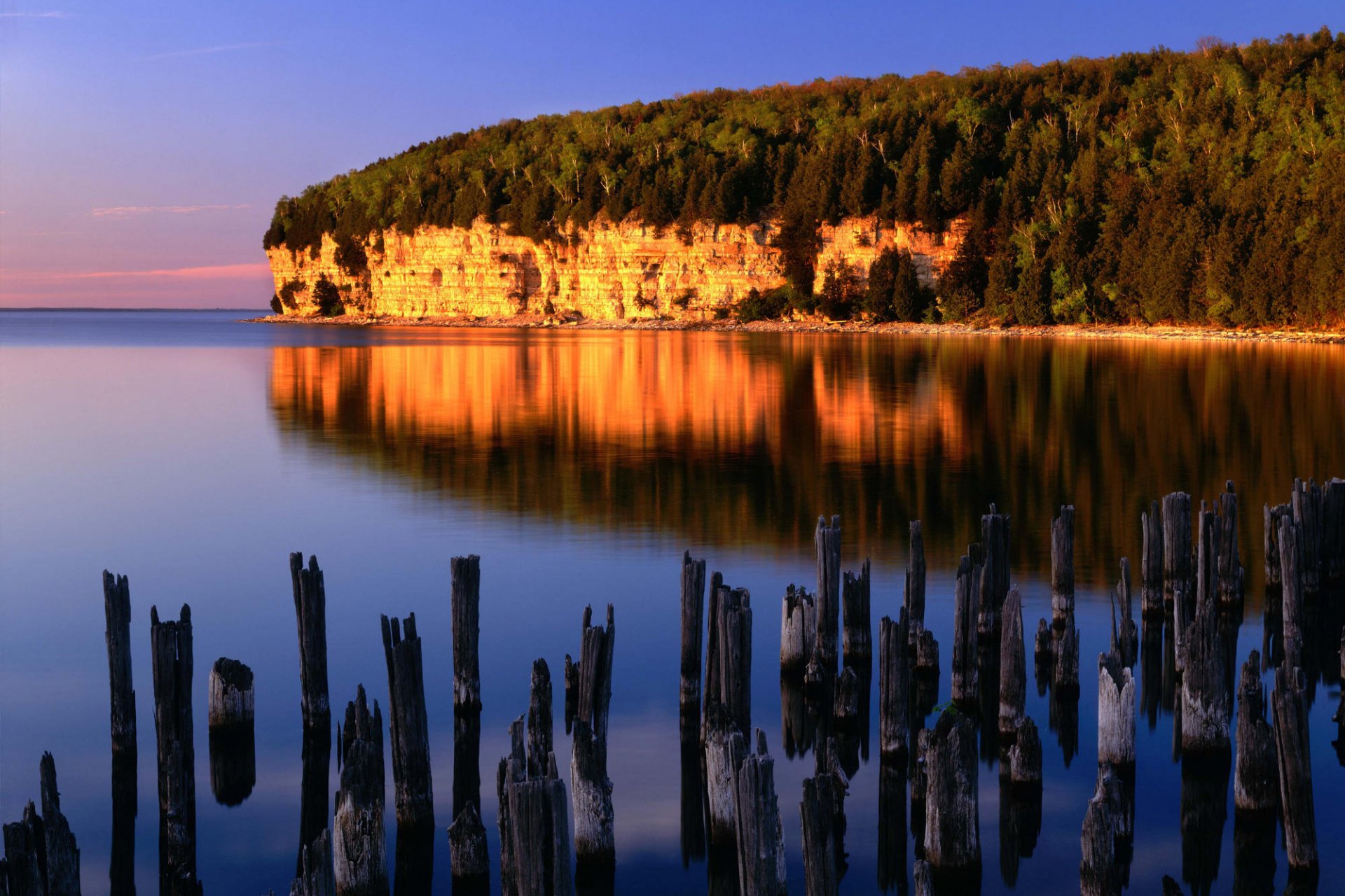 A serene nature scene featuring a cliff reflecting warm colors in calm waters, with weathered stumps in the foreground, creating a stunning HD wallpaper background.