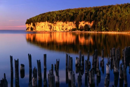 A serene nature scene featuring a cliff reflecting warm colors in calm waters, with weathered stumps in the foreground, creating a stunning HD wallpaper background.