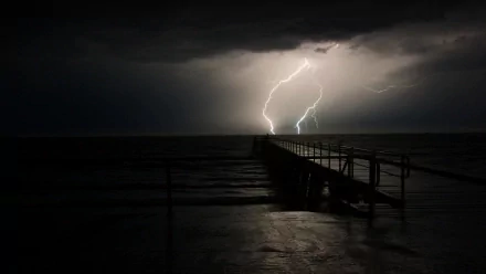 A black and white HD desktop wallpaper featuring dramatic lightning striking near a long pier extending into the dark ocean. The intense light contrasts with the night sky, captured beautifully in this photography.