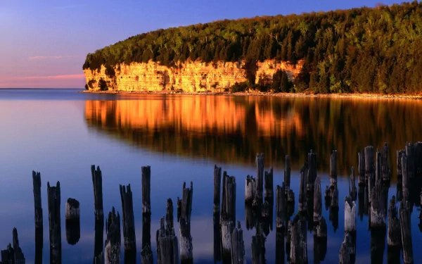 A serene nature scene featuring a cliff reflecting warm colors in calm waters, with weathered stumps in the foreground, creating a stunning HD wallpaper background.