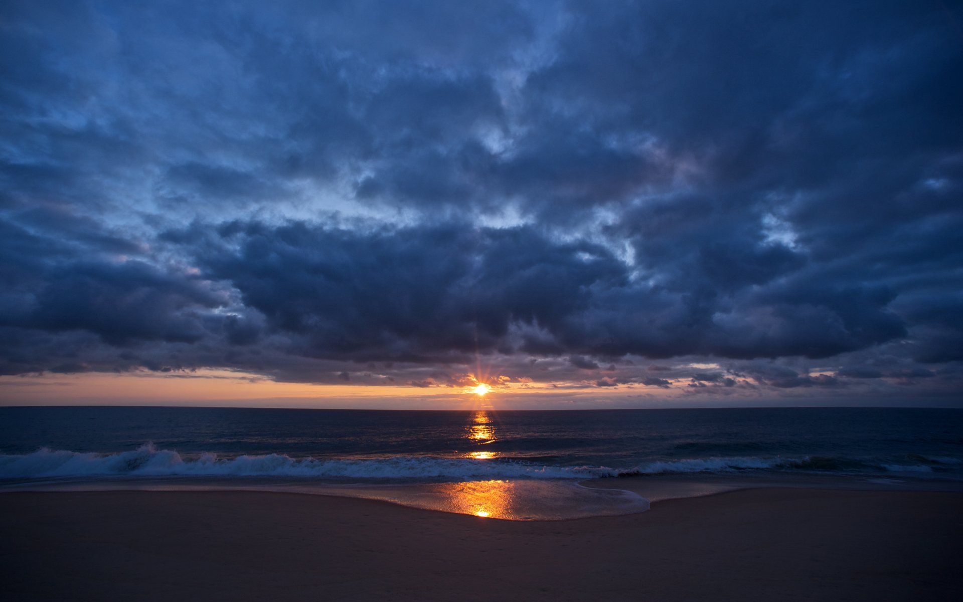 HD desktop wallpaper of a beach at sunrise with ocean waves, dramatic clouds, and natural colors reflecting on the water.