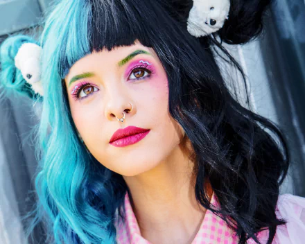 Close-up HD portrait of American singer Melanie Martinez with half blue, half black hair, bold lipstick, and colorful eye makeup, set against a dark background.