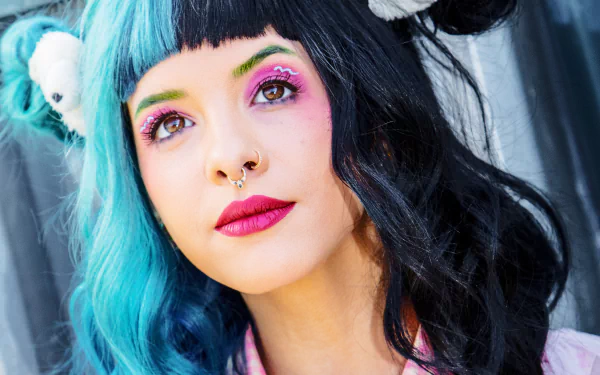 Close-up HD portrait of American singer Melanie Martinez with half blue, half black hair, bold lipstick, and colorful eye makeup, set against a dark background.
