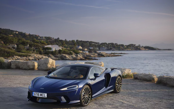 Blue McLaren GT supercar parked on a rocky seaside quay at sunset — sleek McLaren vehicle in high-detail 8K Ultra HD PC desktop wallpaper and background.
