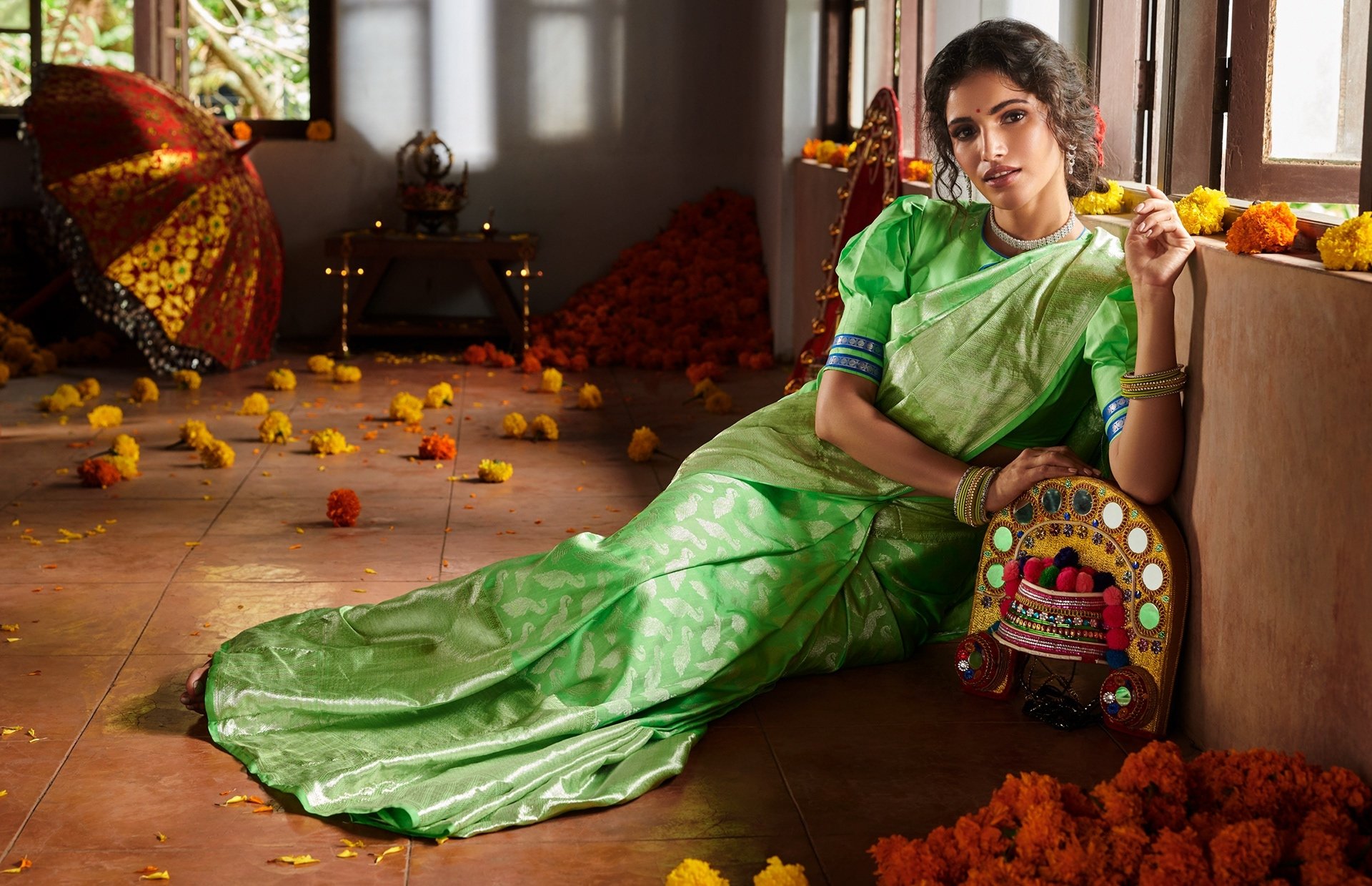 Brunette Indian model Vartika Singh in a vibrant green saree poses gracefully indoors, surrounded by marigold flowers and traditional decor, in this HD desktop wallpaper.