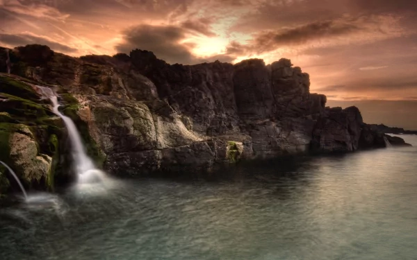 HD desktop wallpaper of Ireland's coastline featuring cliffs, a flowing waterfall and stream, ocean waters, and dramatic cloud-filled sky at sunset.
