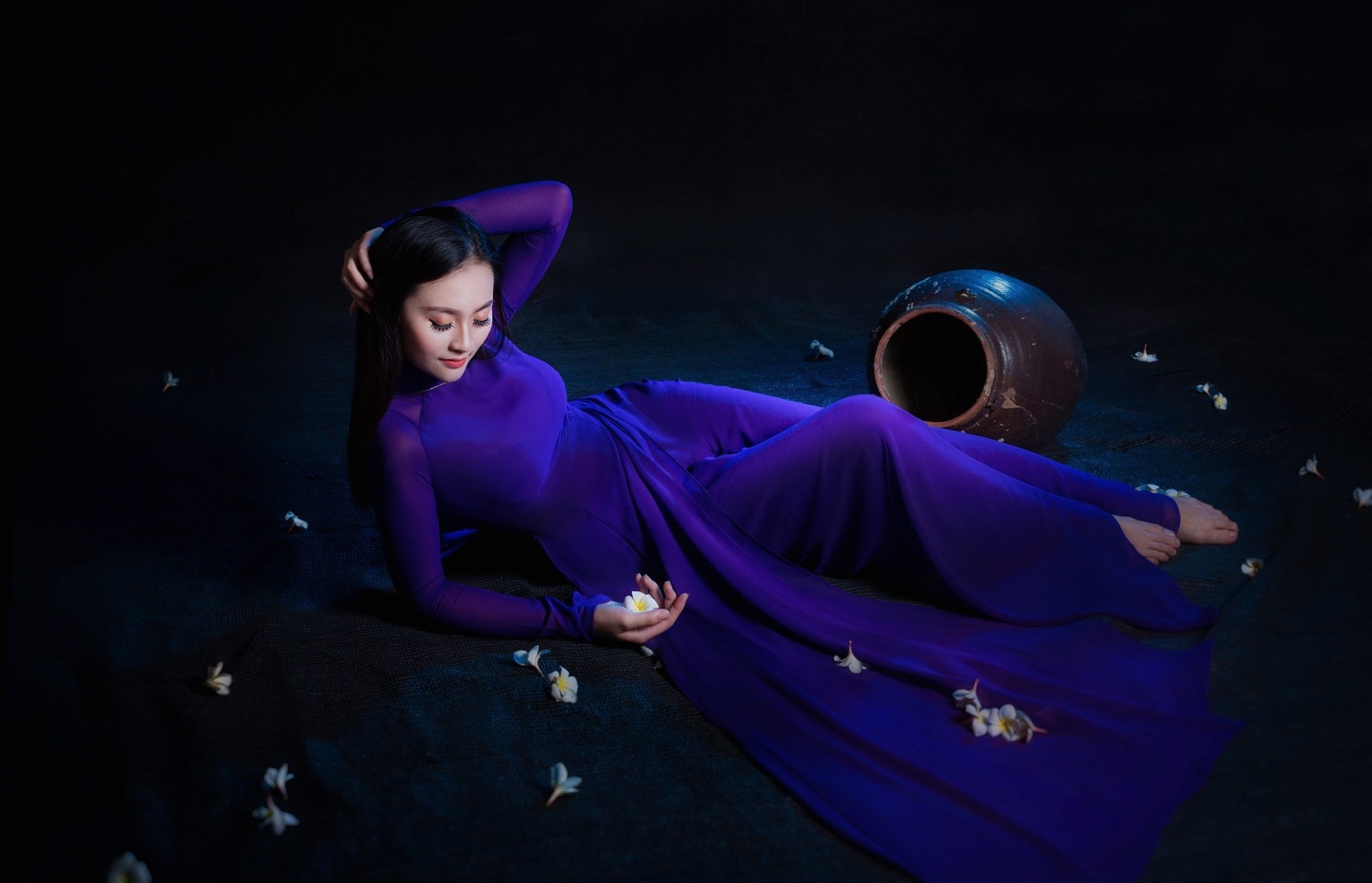 HD wallpaper of an Asian woman with black hair lying down in a flowing purple dress, surrounded by scattered white flowers and a large ceramic vase on a dark background.