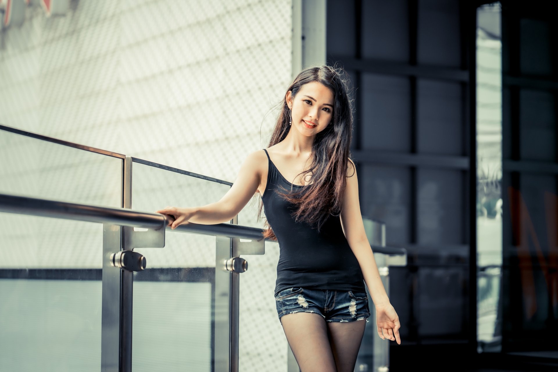 Smiling Asian woman with long black hair wearing a black tank top and shorts, standing by a glass railing in a modern setting, captured in 4K Ultra HD resolution.