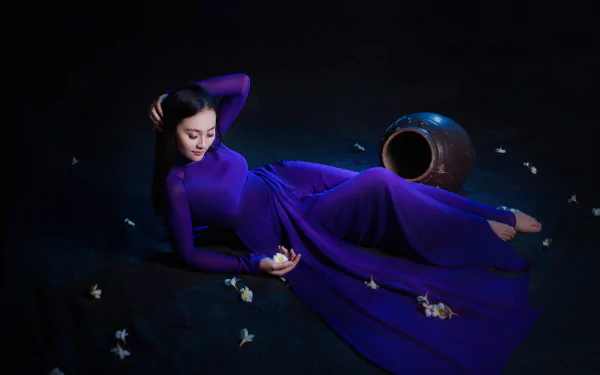 HD wallpaper of an Asian woman with black hair lying down in a flowing purple dress, surrounded by scattered white flowers and a large ceramic vase on a dark background.