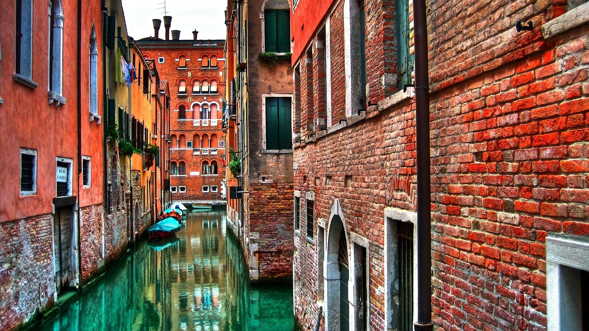 An HD desktop wallpaper featuring a picturesque canal in Venice, Italy, bordered by colorful, rustic buildings and boats on the emerald-green water.