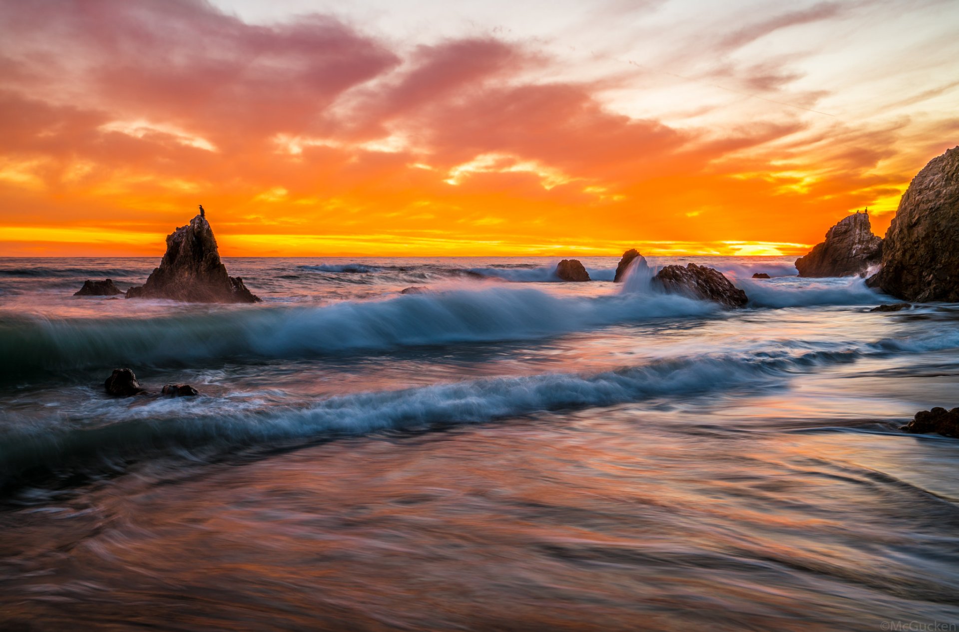 HD PC desktop wallpaper of ocean waves at sunset, golden-orange sky over a rocky shore with rolling waves and reflective wet sand.