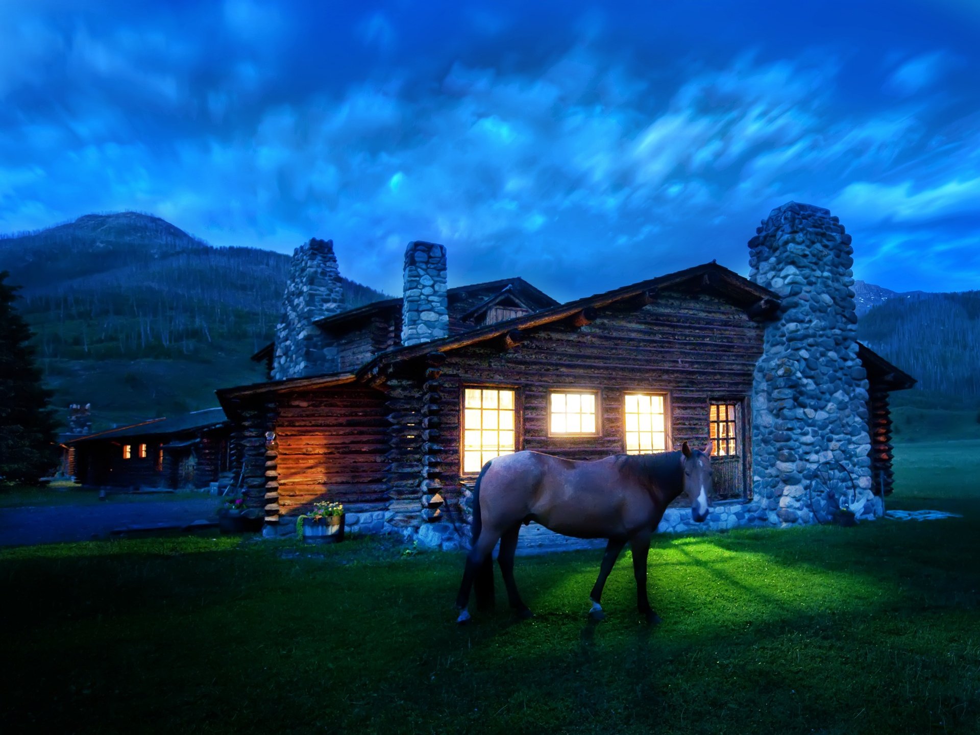 HD desktop wallpaper featuring a horse standing on a green lawn in front of a rustic log cabin with lit windows, set against a dramatic evening sky and scenic mountain backdrop.
