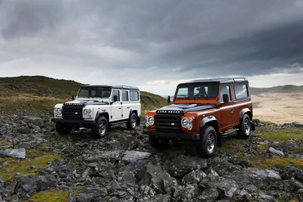 HD wallpaper featuring a rugged landscape with an orange and a white Land Rover Defender SUV. The vehicles are showcased against a dramatic sky, emphasizing their off-road capabilities.