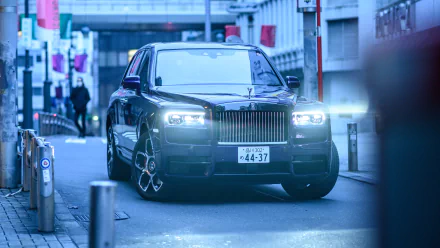 5K Ultra HD PC desktop wallpaper and background of a black Rolls-Royce Cullinan vehicle on an urban street at dusk, headlights on, Spirit of Ecstasy hood ornament and grille in view.