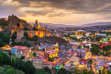 A stunning HD desktop wallpaper showcasing a vibrant cityscape of Georgia, bathed in warm light during sunset with historic buildings and surrounding hills in view.