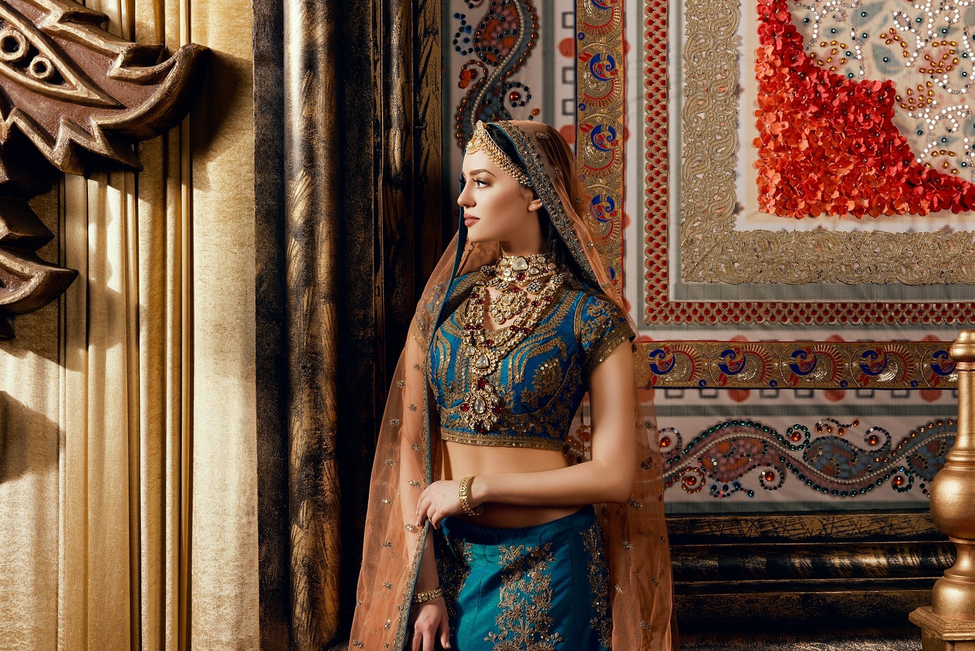 HD PC desktop wallpaper of an Indian woman model in a blue embroidered sari with ornate necklace, posed against a richly decorated palace-style background.