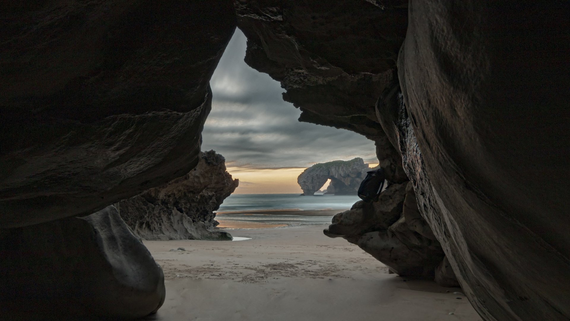2K Quad HD PC desktop wallpaper: a nature scene viewed from inside a dark rocky cave opening onto a sandy beach with a distant sea arch at sunset beneath a cloudy sky.