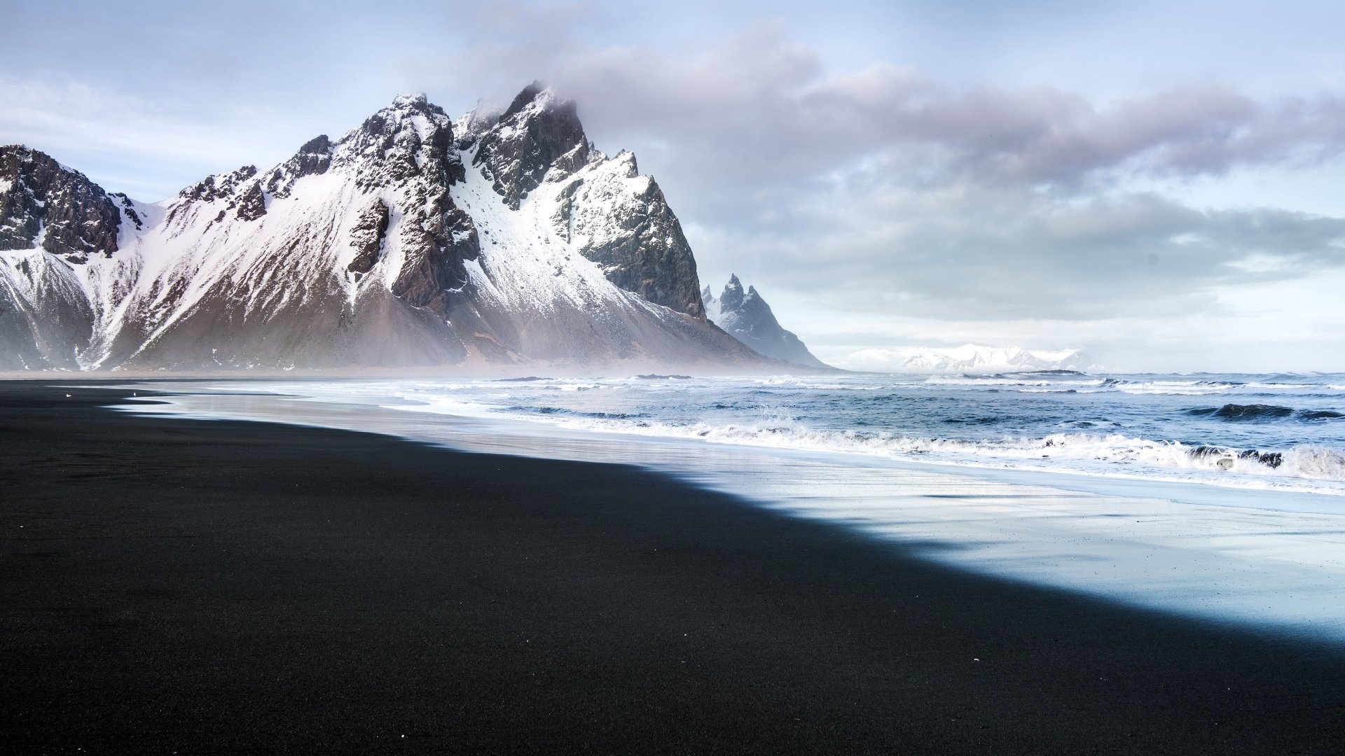 Snow-capped Vestrahorn mountains beside a black sand beach with waves under a cloudy sky, captured in stunning 4K Ultra HD for a nature-themed desktop wallpaper.
