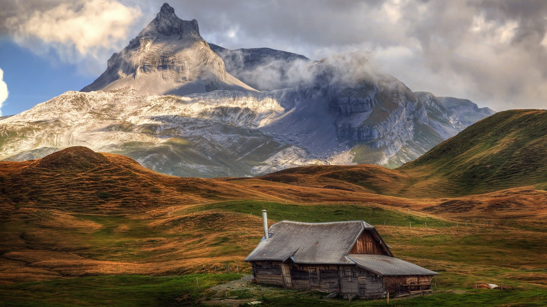 4K Ultra HD PC wallpaper of a man-made cabin set against a dramatic mountain backdrop with rolling hills under a partly cloudy sky.