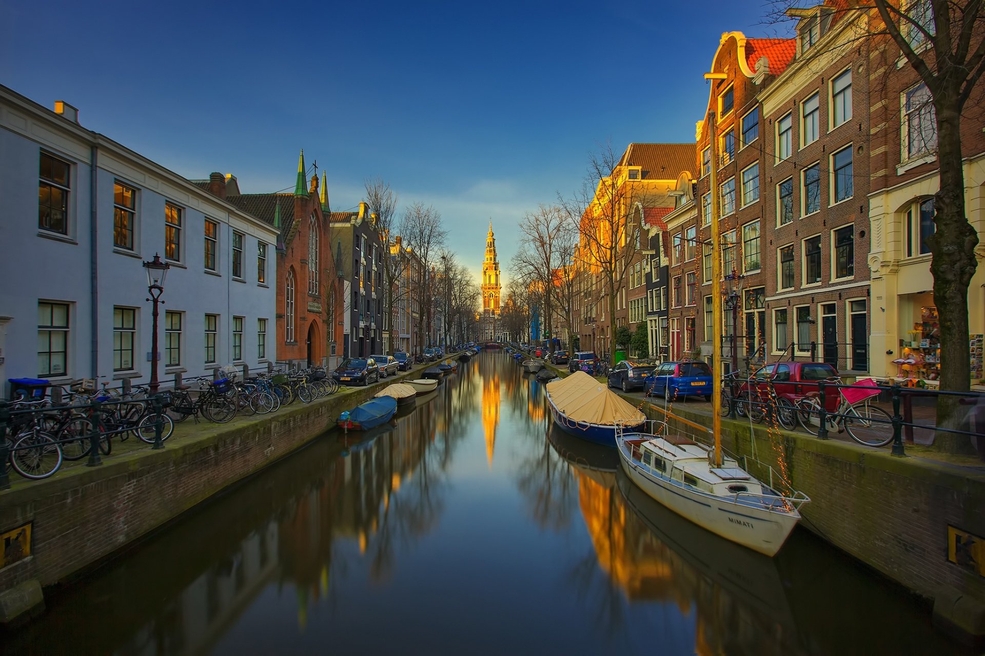 HD desktop wallpaper of a man-made canal in Amsterdam, featuring historic buildings and boats under a clear blue sky at sunset.