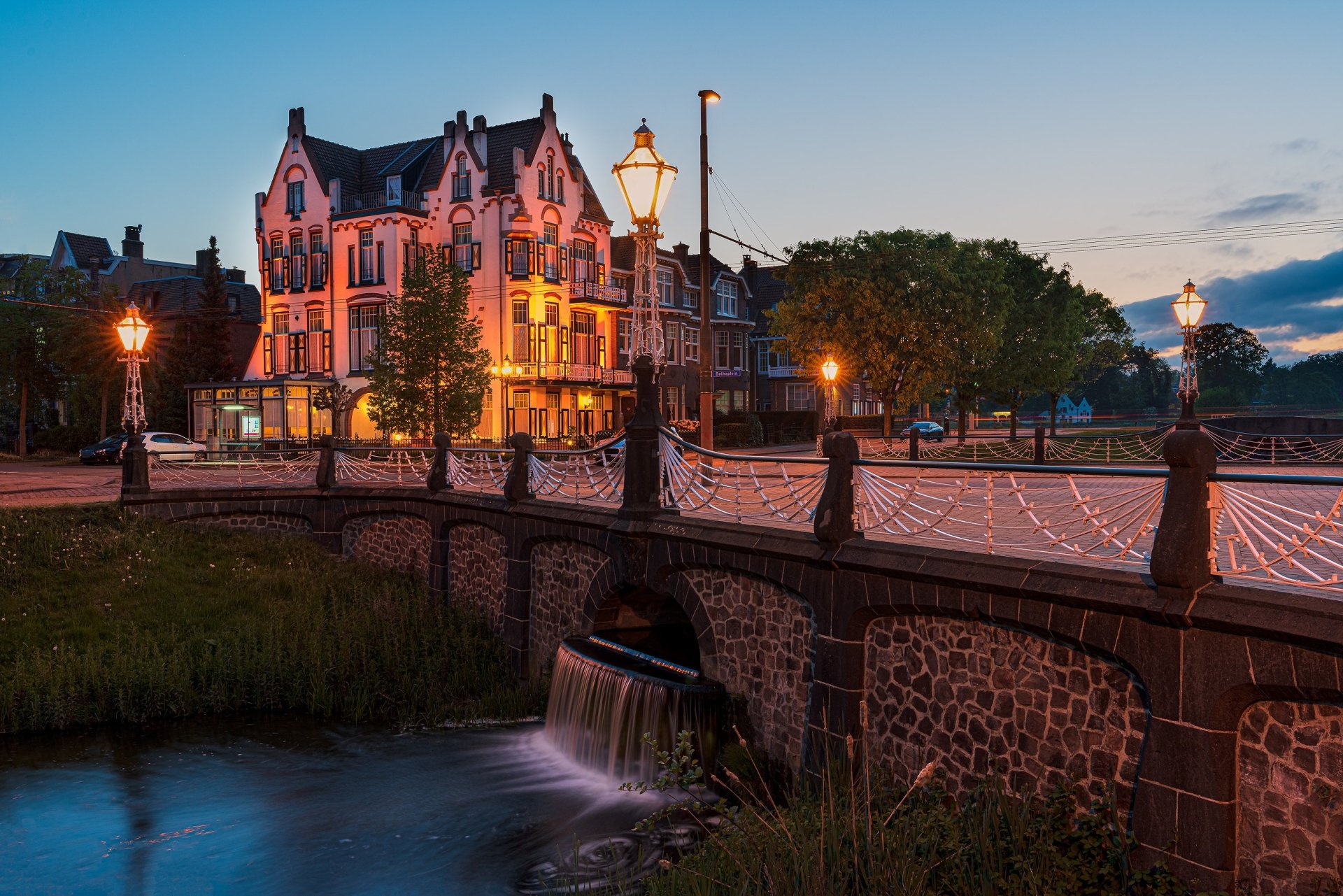 2K desktop wallpaper showing an evening scene in Arnhem, Netherlands: a lamp-lit hotel beside a river and man-made stone bridge.