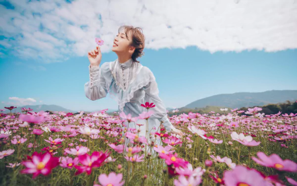 8K Ultra HD PC desktop wallpaper of a brunette model in a field of pink flowers, dreamy mood with shallow depth of field and soft bokeh as she smiles and reaches toward the sky.