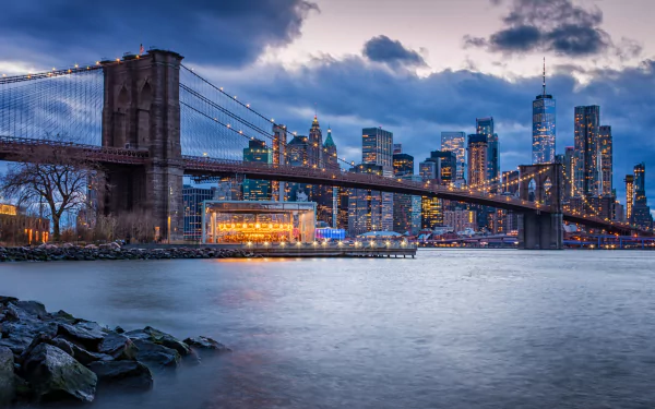 4K Ultra HD view of the Brooklyn Bridge spanning the East River with New York City skyscrapers in the background under a dramatic sky.