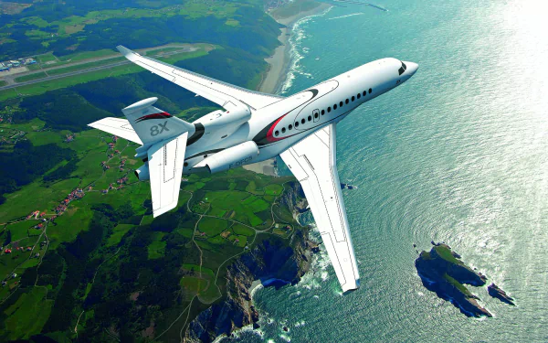 A Dassault Falcon 8X private jet aircraft soars over a coastal landscape, with green fields and ocean below, displayed in HD as a desktop wallpaper and background.