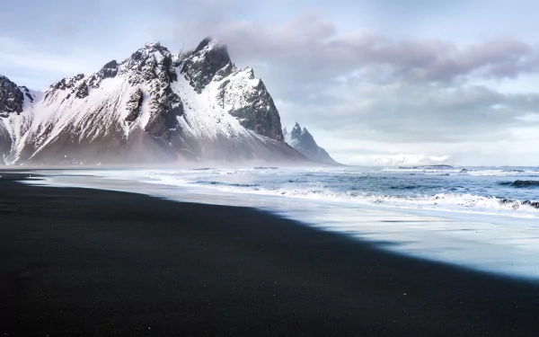 Snow-capped Vestrahorn mountains beside a black sand beach with waves under a cloudy sky, captured in stunning 4K Ultra HD for a nature-themed desktop wallpaper.