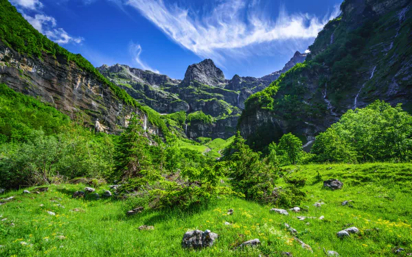 A vibrant 4K Ultra HD desktop wallpaper showcasing lush green mountains with rugged stone cliffs under a bright blue sky with wispy clouds.