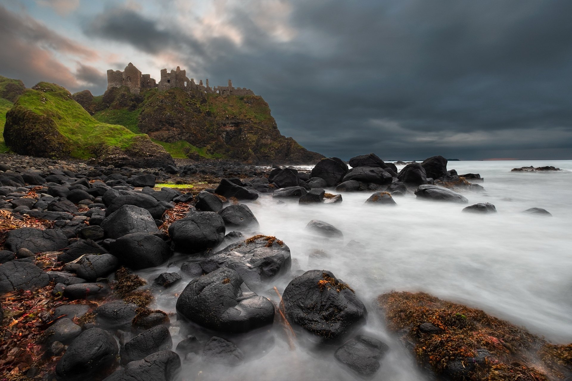 Dunluce Castle stone ruins perched on a rocky coastline in Ireland, under a moody sky with waves crashing against dark stones.