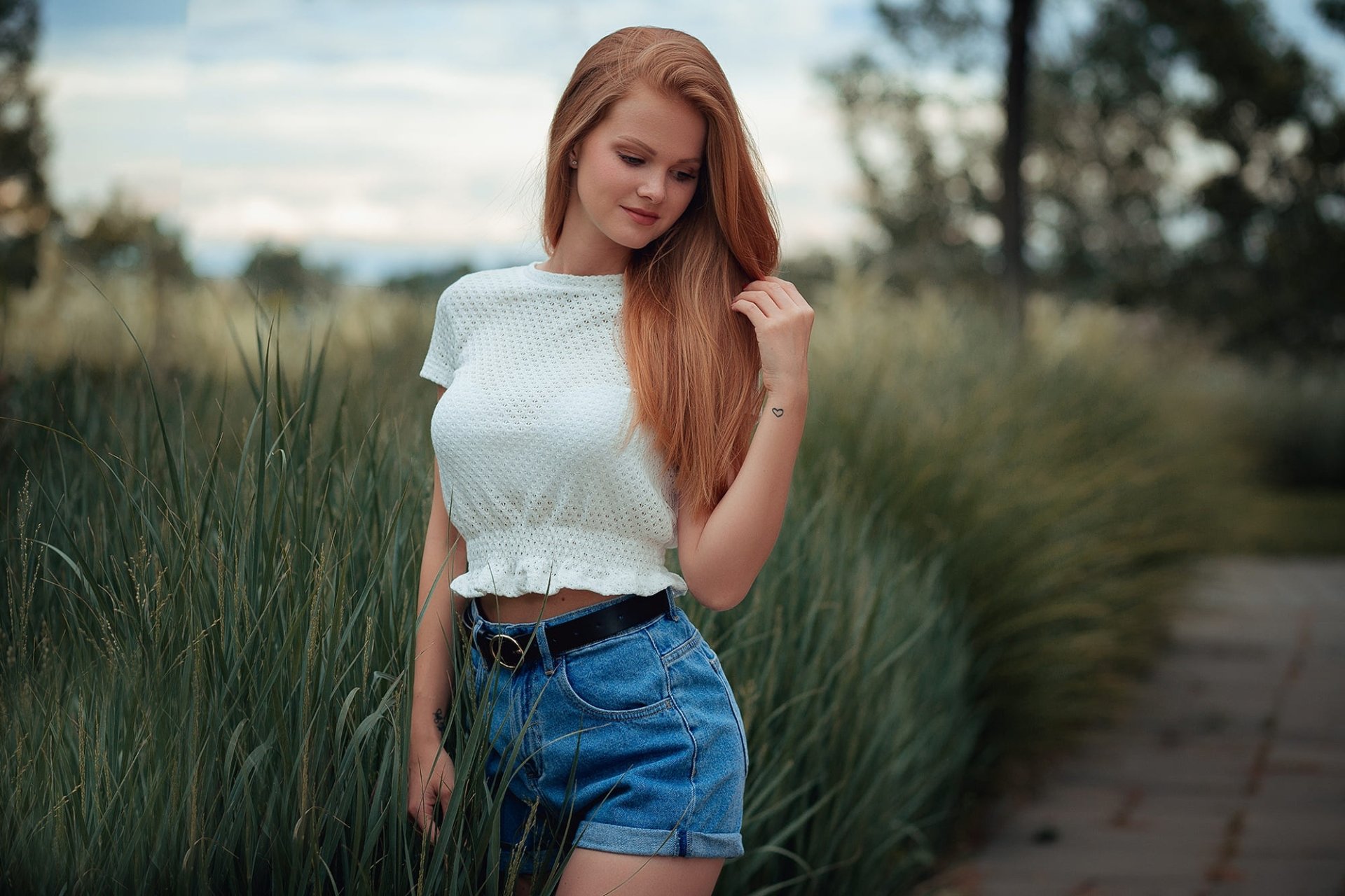 Redheaded female model in denim shorts and a white cropped top, posed amid tall grass with soft depth of field — HD desktop wallpaper/background.