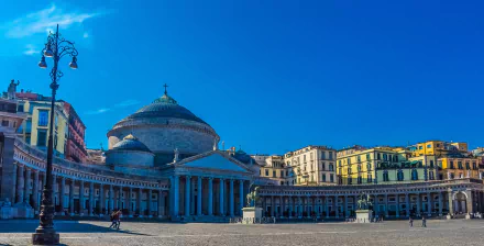 Town square in Naples, Italy, featuring a prominent religious church with classical architecture and statues under a clear blue sky.